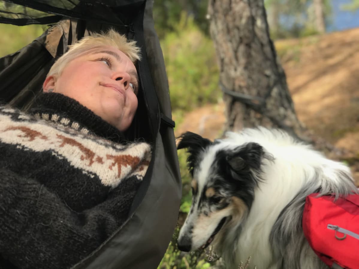 Riippumatossa makaava nainen hymyilee koiralle, joka istuu vieressä