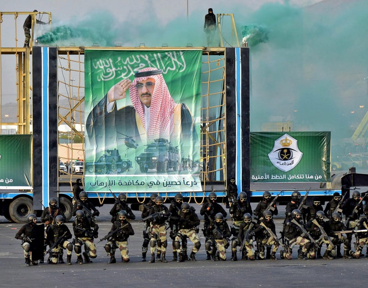 Saudien erikoisjoukkoja on kahdessa rivissä suuren julisteen edessä. Sotilailla on kädessään rynnäkkökivääreitä, yllään mustat paidat, maastokuvioidut housut, kypärät, suojalasit ja kasvonaamiot. Julisteessa taustalla on kuva prinssi Mohammed bin Naif Abdulazizista, joka on maan sisäministeri.