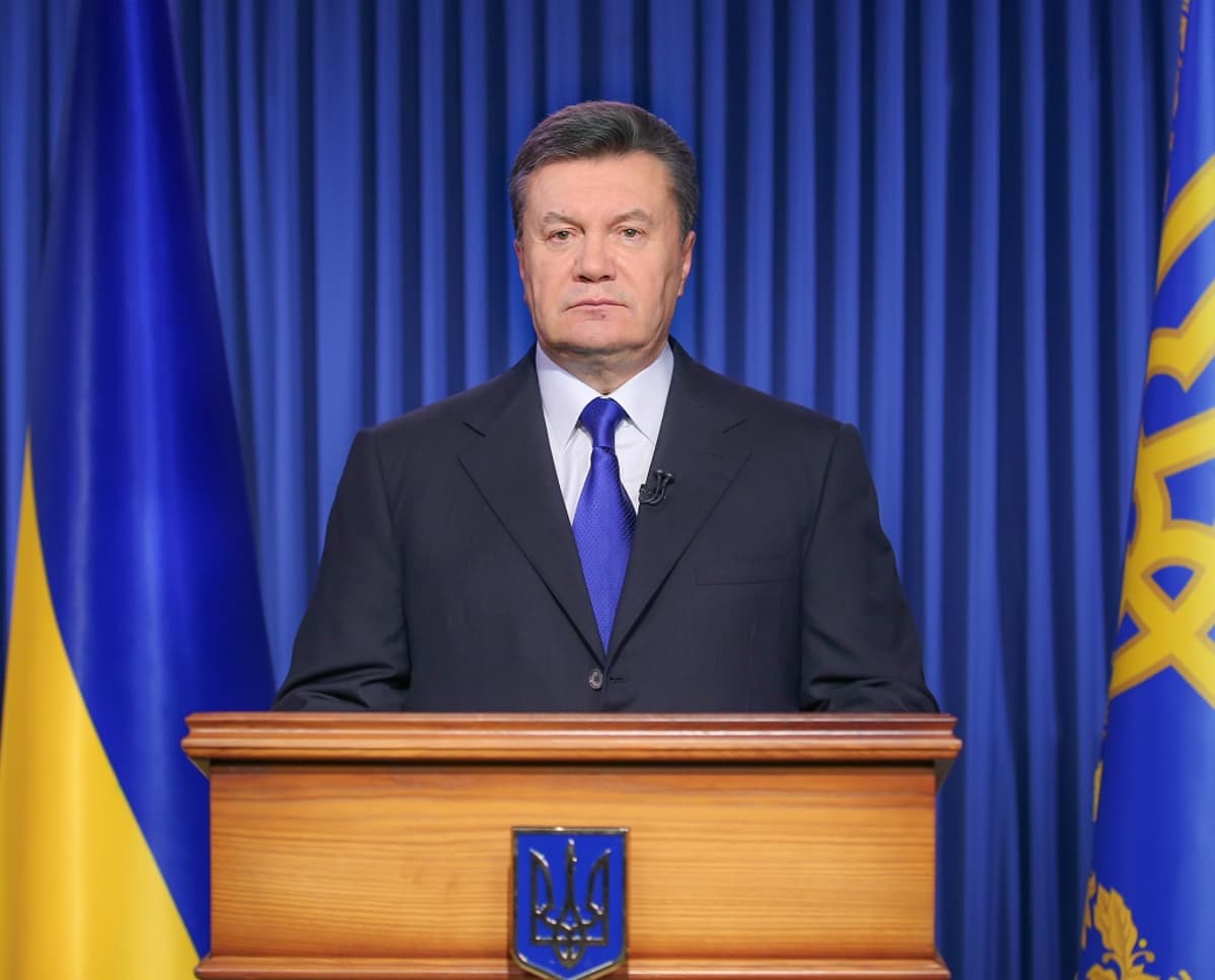 Janukovytš seisoo vakavana korokkeen takana tummassa puvussa. Taustalla on sininen verho ja Ukrainan lippu. Janukovytšin kravatti on samaa väriä.