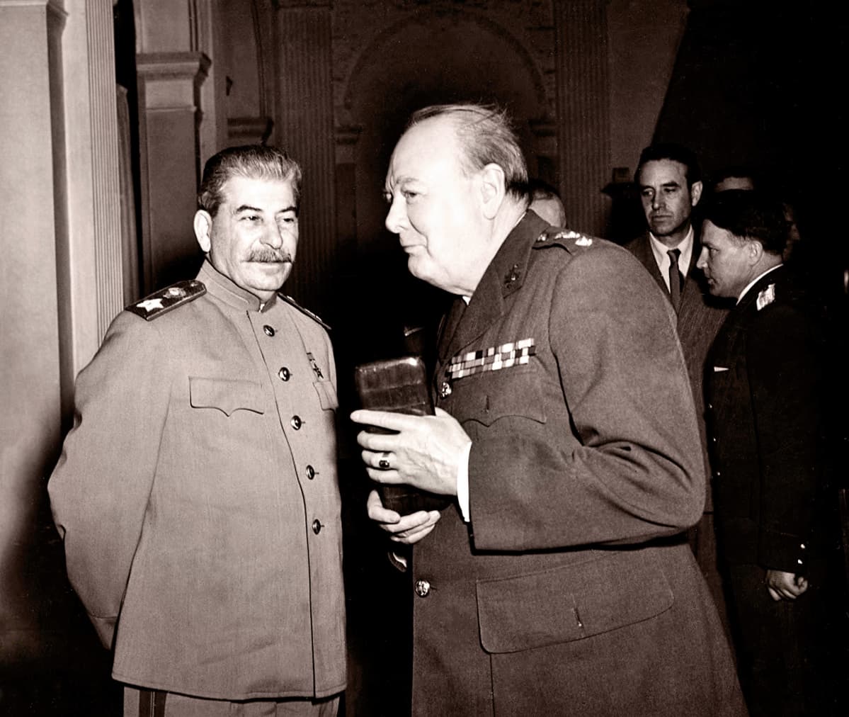 Stalin ja Churchill seisovat vierekkäin, taustalla näkyy muita miehiä. Molemmilla on univormut. Stalin katsoo Churchilliä, Churchill katsoo toisaalle.