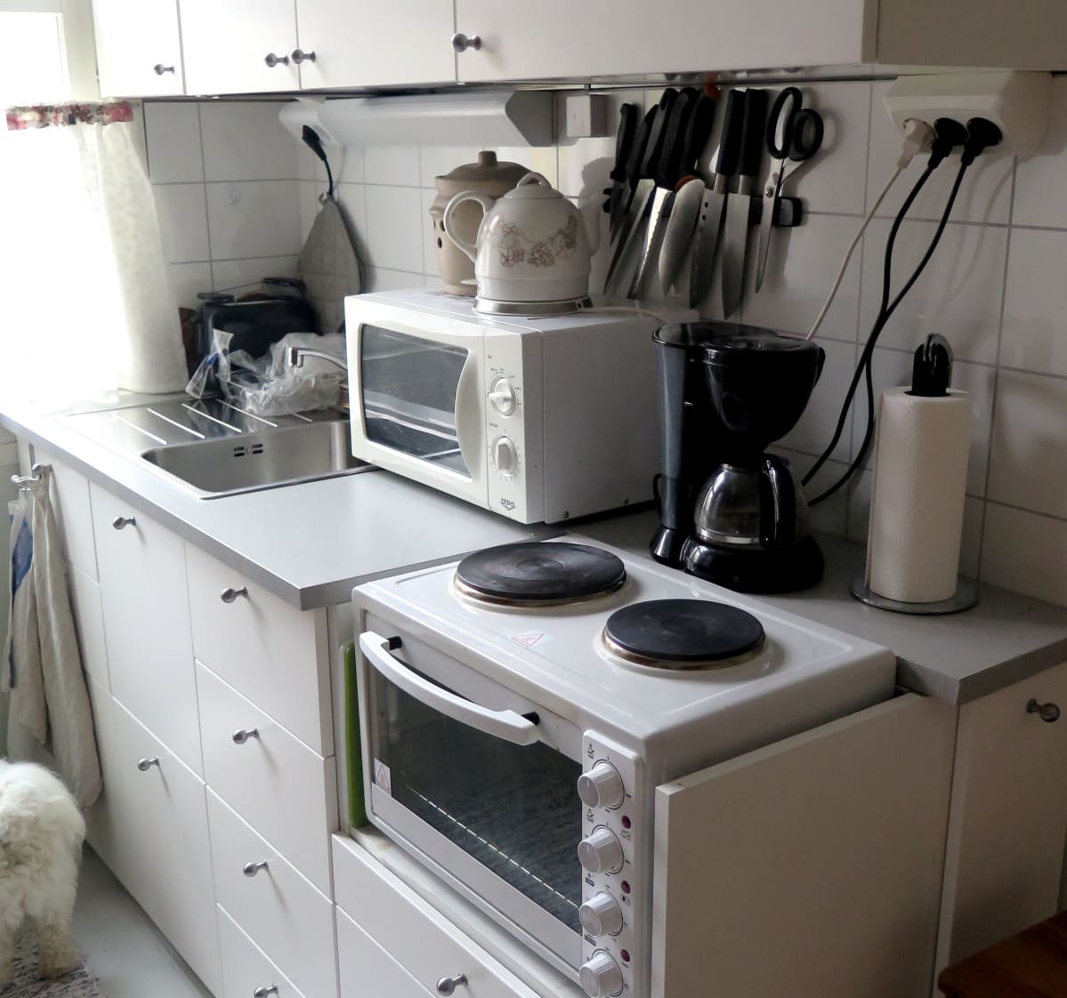 Valkoinen, pieni keittiö. Kaappeja. Työpöytätaso. Mikroaaltouuni ja hella.