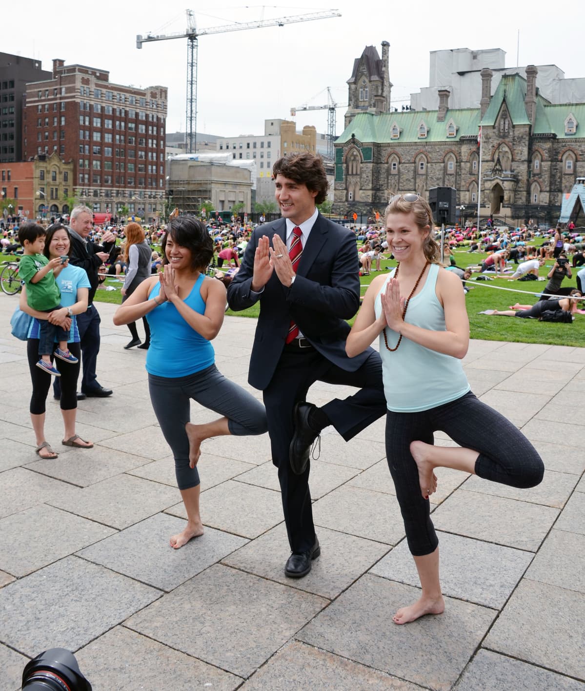 Tummaan pukuun pukeutunut Justin Trudeau koukistaa polveaan jooga-asentoon, vierellä kaksi voimisteluasuista naista tekee samaa.