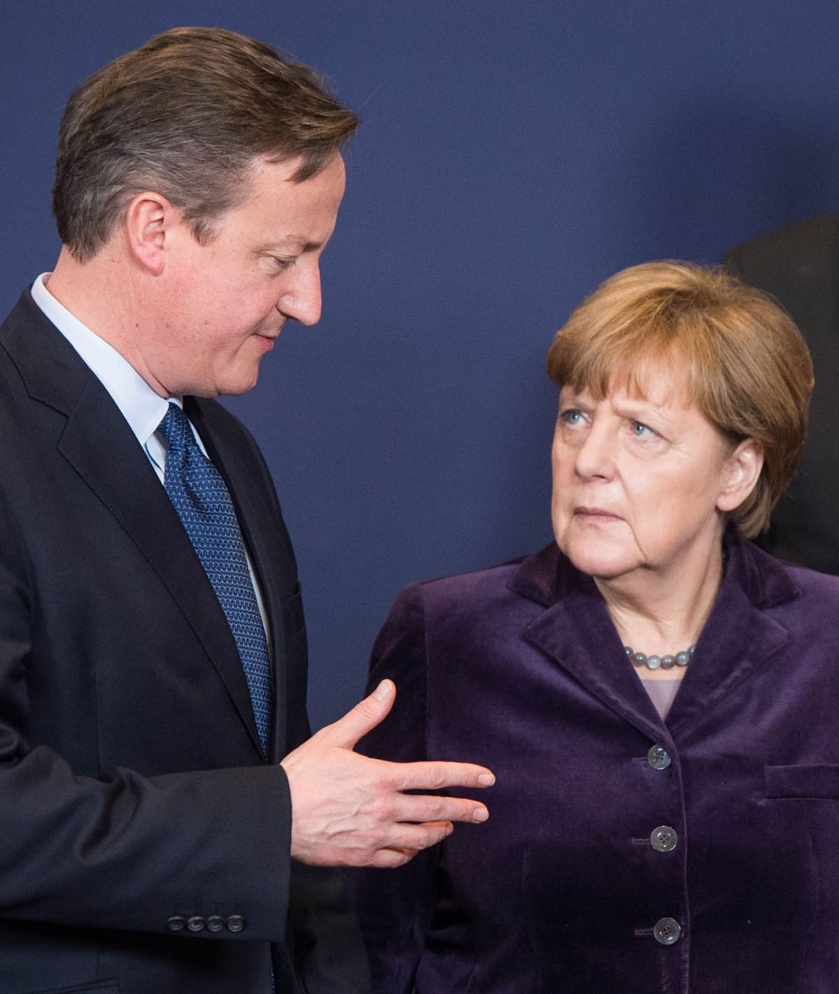 Britannian pääministeri David Cameron selittää jotain Angela Merkelille. Hänen oikea kätensä on ojennettuna. Cameronin kasvoilla on vieno hymy. Merkel katsoo Cameronia vakavana, ehkä jopa hieman huolestuneen oloisena. Cameronilla on tumma puku, sininen pilkullinen kravatti, Merkelillä tummanvioletti asu. Tausta on tummansininen.
