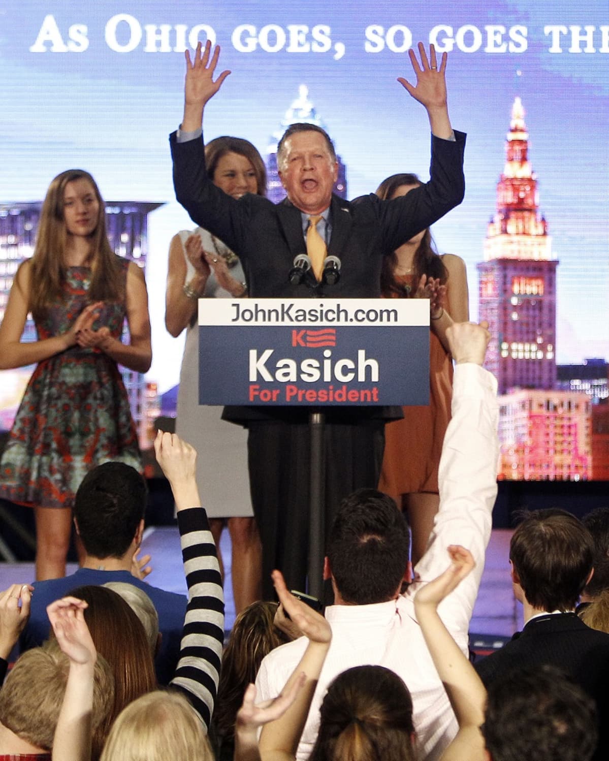 Kasich kädet ylhäällä puhujapöntössä, taustalla vaimo ja kaksi tytärtä.