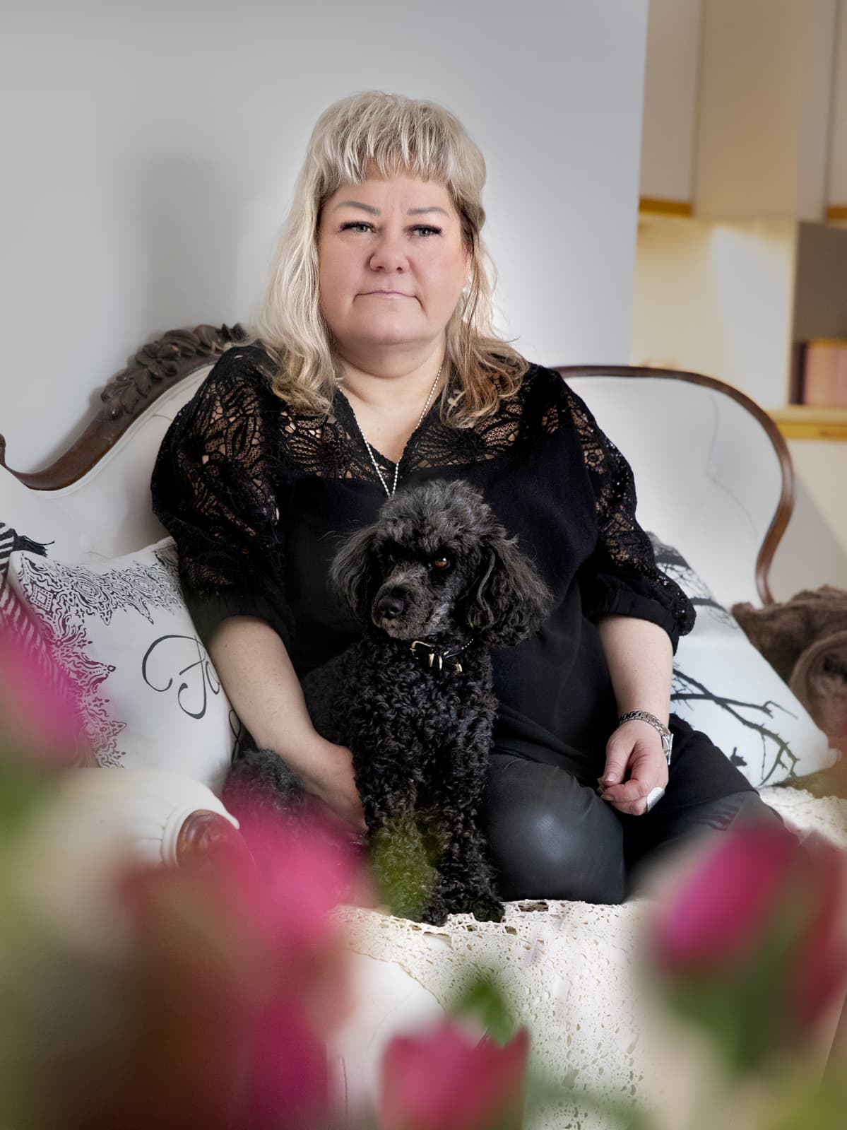 Maarit Pitkänen istuu kotonaan valkoisella sohvalla sylissään musta pieni villakoira Leenu. Etualalla ruusuja.