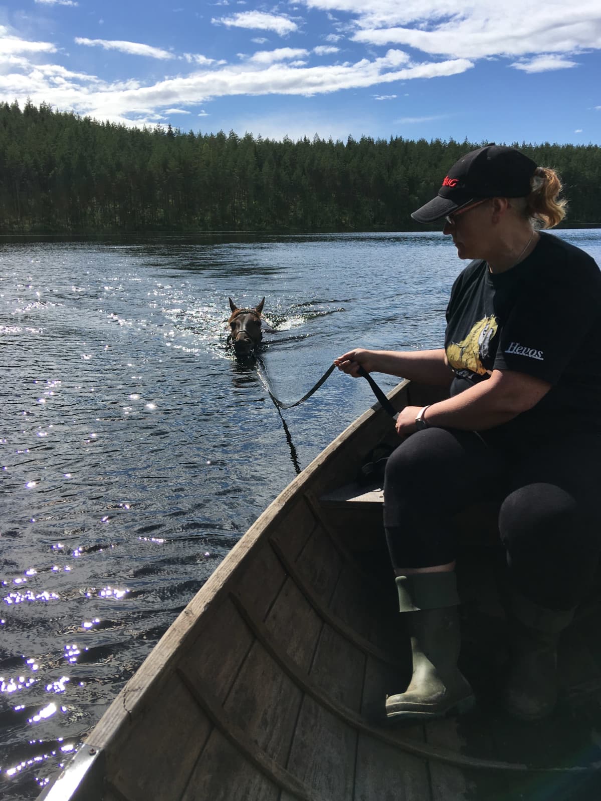 Nainen istuu veneessä, hevonen ui vedessä veneen perässä