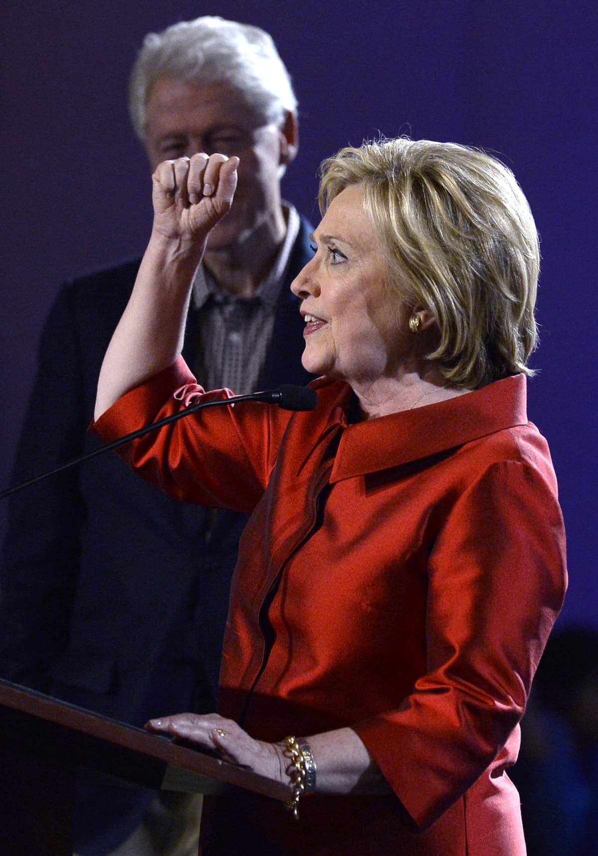 Hymyilevä Hillary Clinton pitää oikeaa kättään pystyssä ja nyrkissä. Taustalla näkyy hänen miehensä Bill, jonka kasvot jäävät nyrkin taakse.