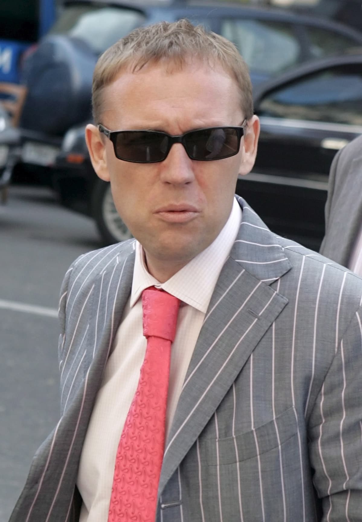 Andrei Lugovoi mustat lasit pääsään, raidallisessa harmaassa puvussa, vaaleanpunainen kravatti kaulassaan.