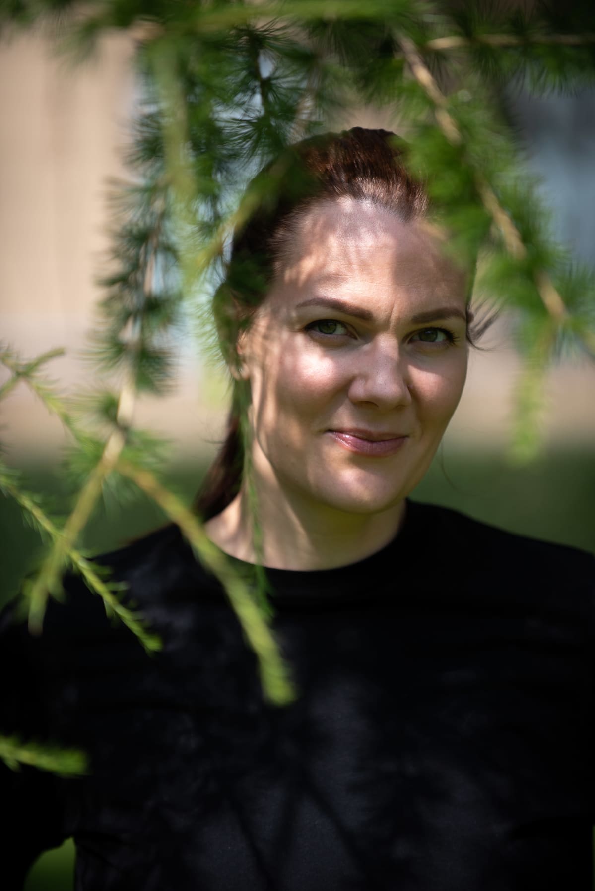 Outi Ellilä seisomassa puistossa, vihreät oksat kehystävät kasvoja.