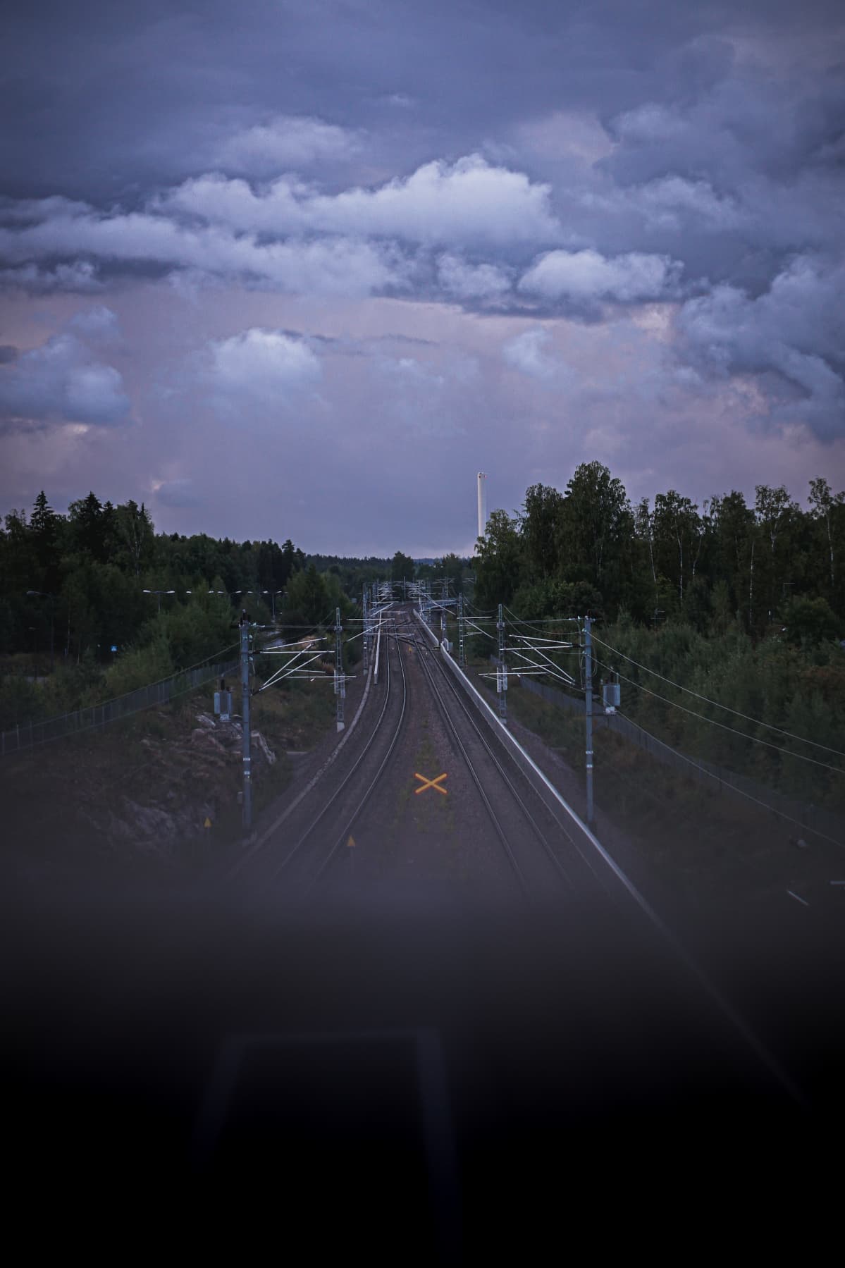 Maisemakuva joka on otettu sillalta. Kuvan keskellä jatkuvat junaraiteet kohti horisonttia. Taivaalla on synkkiä, tumman purppuraisia pilviä.