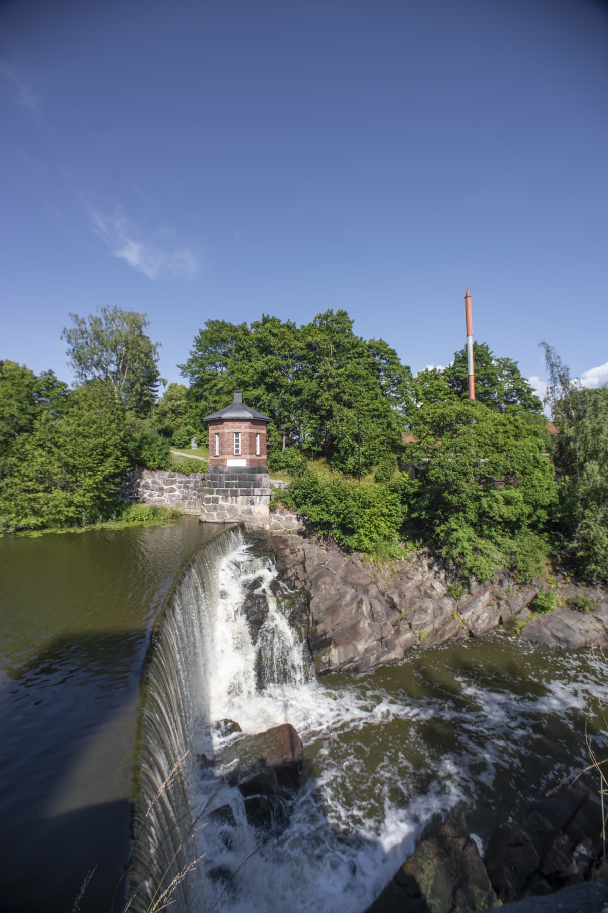 En damm som vattnet forsar över, ner i ett vattenfall. I bakgrunden syns träd och ett litet tegelhus. Det är en solig dag, himlen är blå.