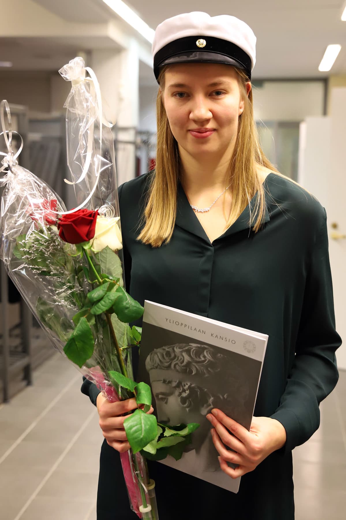Mette Baas valkolakki päässään, kukat ja ylioppilastutkintotodistus kädessään.