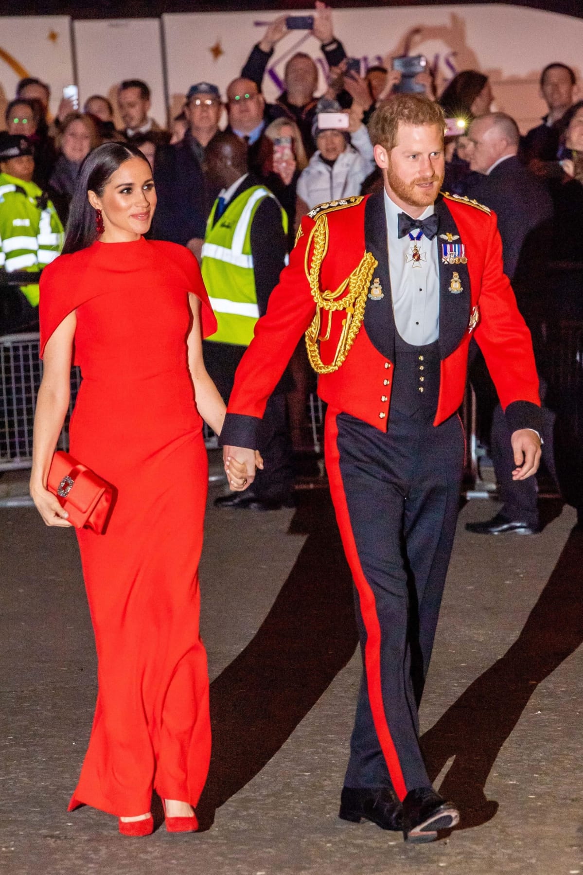 Punaisessa iltapuvussa oleva Meghan ja univormussa oleva Harry pitävät toisiaan kädestä.