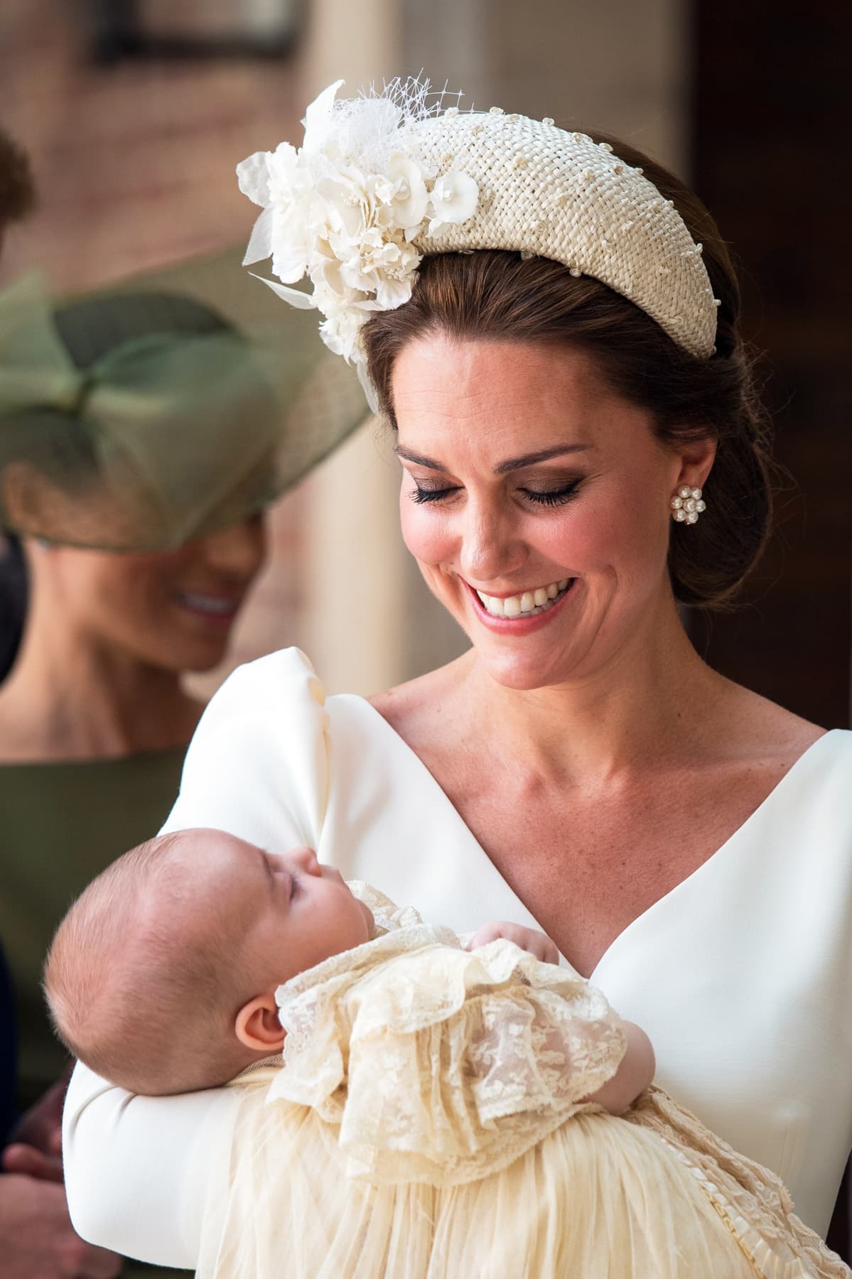 Cambridgen herttuatar Catherine sylissään prinssi Louis kastemekossaan. Taustalla näkyy Sussexin herttuatar Meghan.