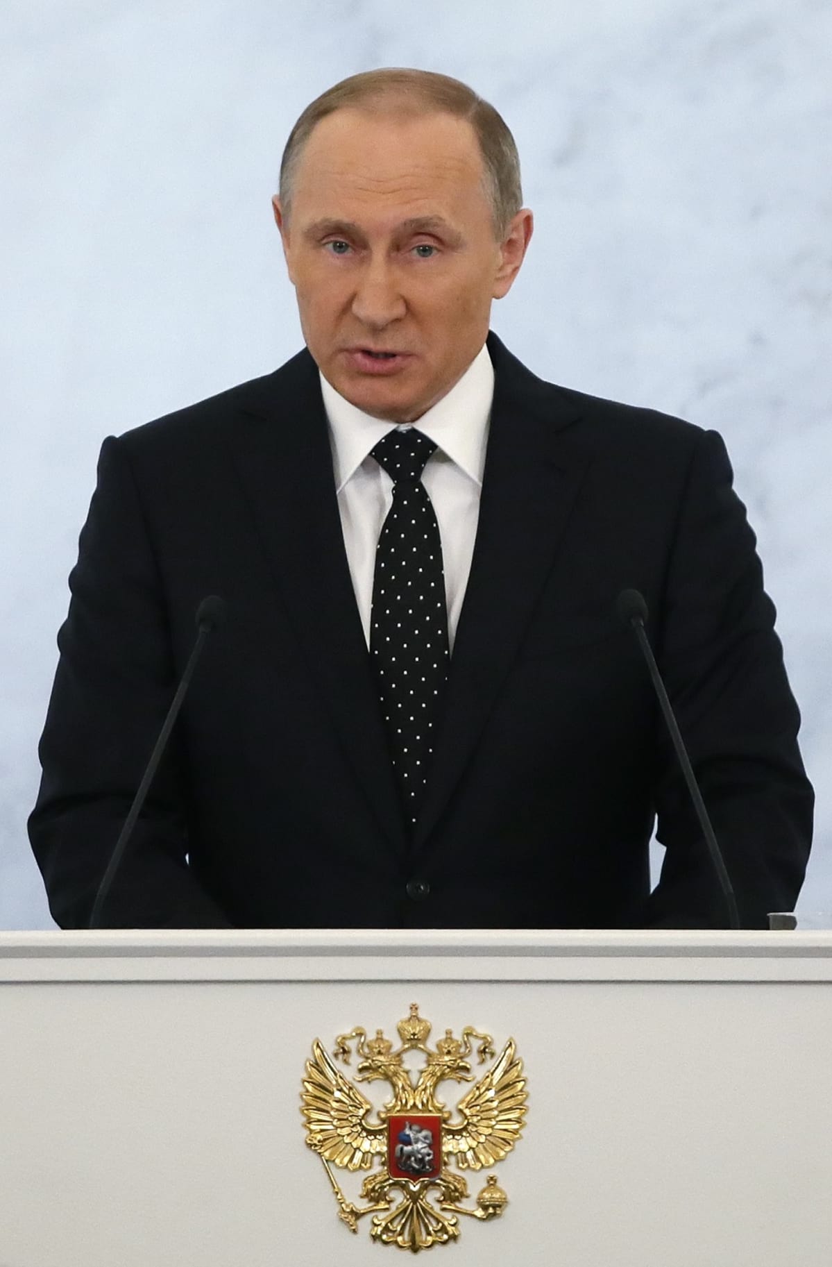 Kuva Venäjän presidentistä puhuu. Hänen edessään on kaksi mikrofonia. Putinilla on musta puku ja musta valkopilkkuinen kravatti. Puhujankorokkeessa on Venäjän vaakunatunnus, kaksipäinen kotka. Tausta on vaalea.