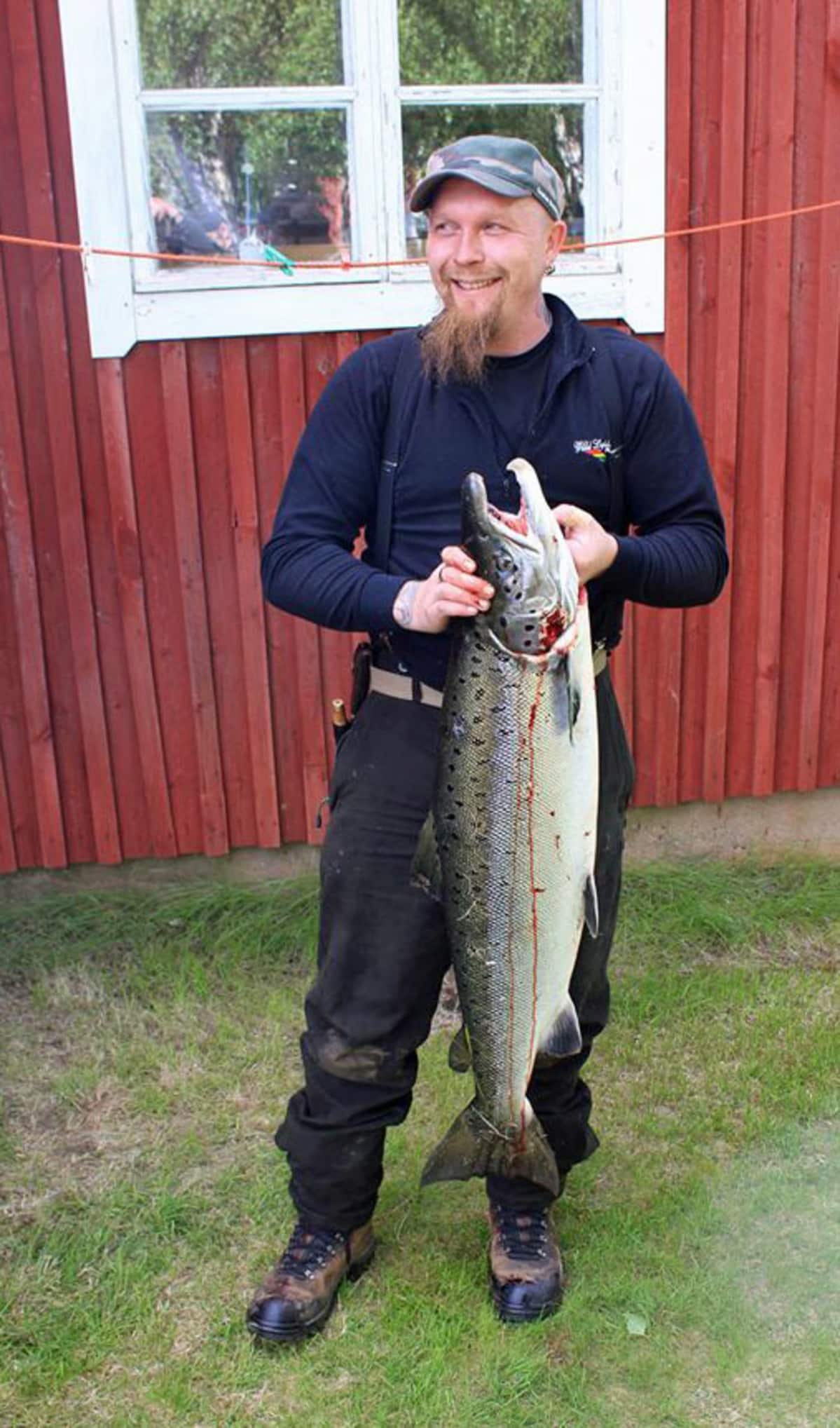 Mies seisoo talon edessä suuri kala käsissään.