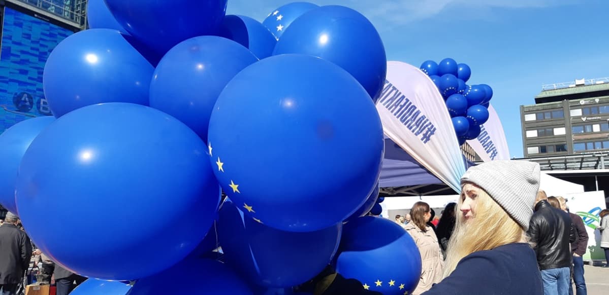 Kuvassa EU-tunnuksilla kuvioituja ilmapalloja kädessään pitävä vaaleahiuksinen nainen.