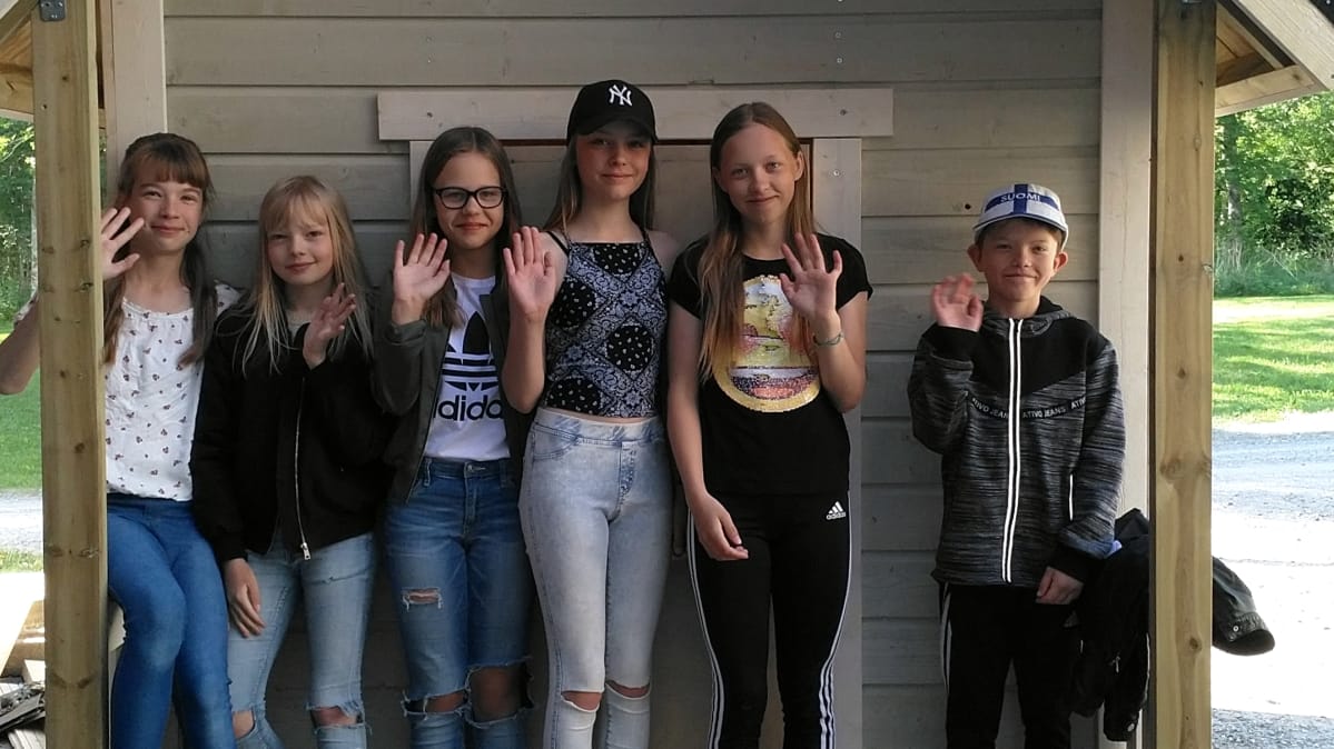 Kuvassa kuusi oppilasta seisoo leikkimökin edessä ja vilkuttaa.