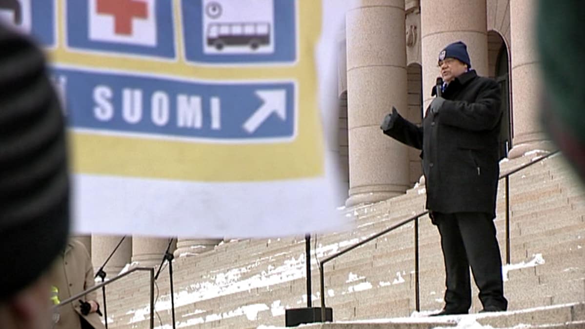Timo Soini pitää puhetta eduskuntatalon portailla helmikuun lopussa 2011, etualalla liehuu perussuomalaisten vaalibanderolli.