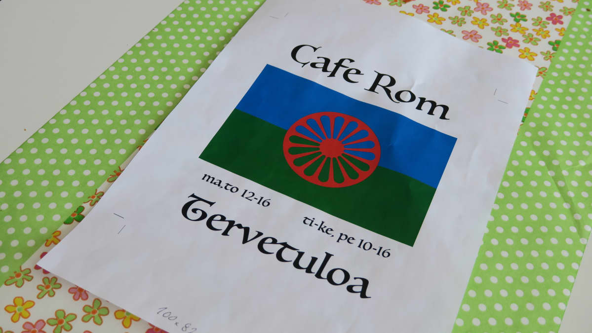 A4-mainoslappu pöydällä. Cafe Rom romanikahvilan mainoksessa romanien lippu.