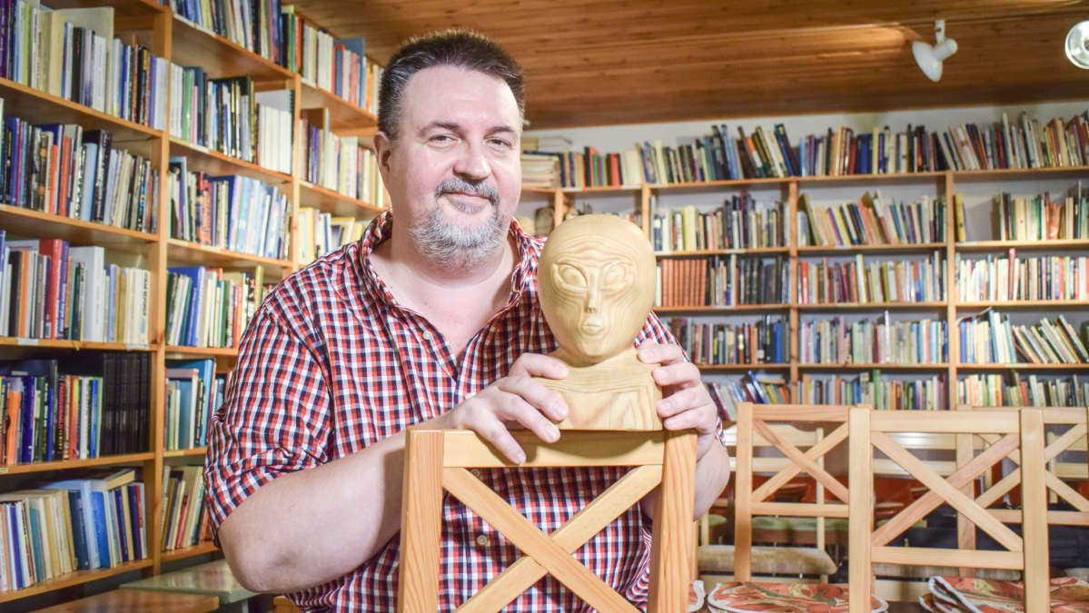 Marko Kananen istuu puisen humanoidipatsaan kanssa kirjahyllyn edessä.