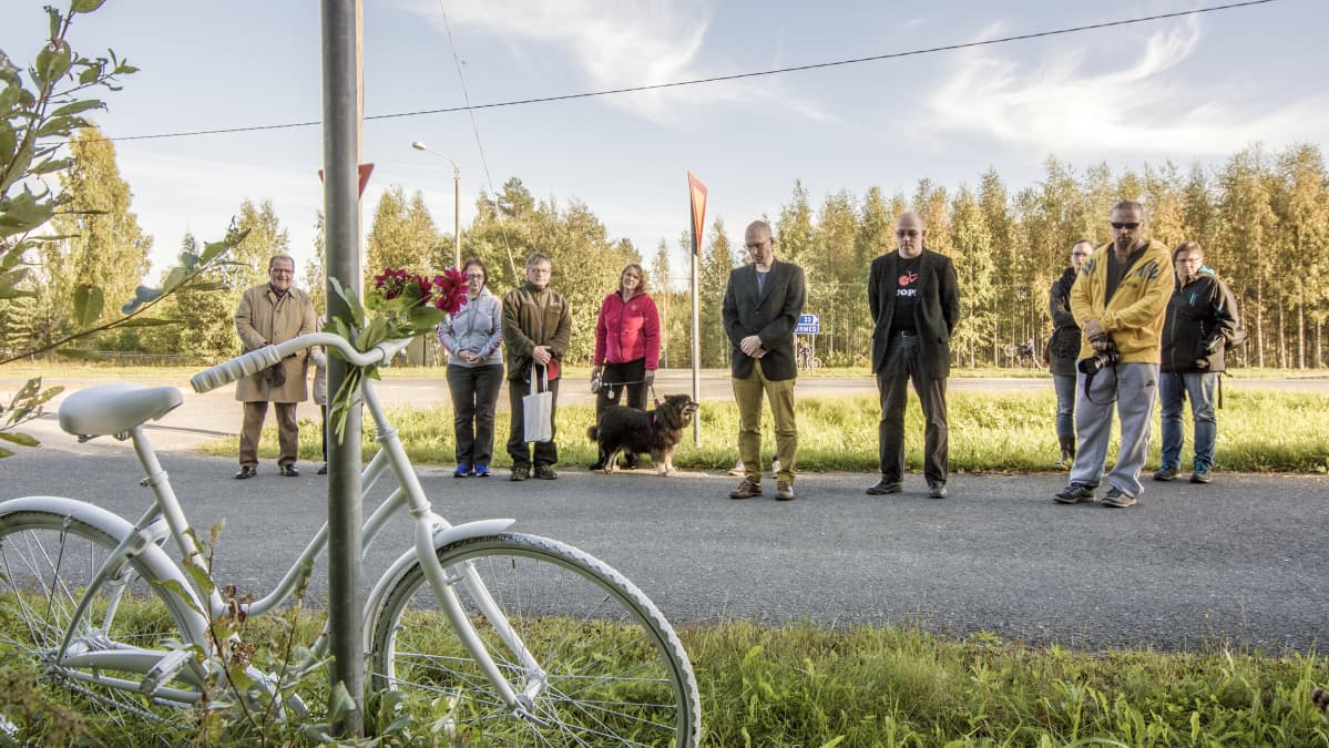 Haamupyörä nojaa liikennemerkkiin. Ihmisiä seisoo taustalla.