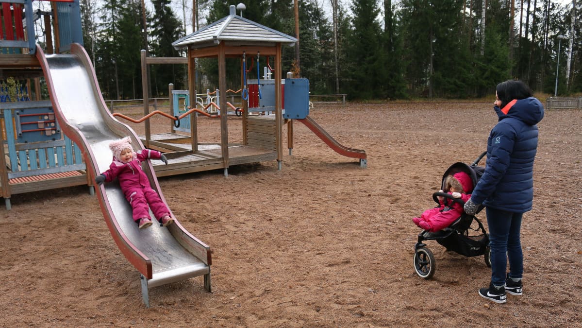 äiti ja kaksi lasta leikkipuistossa, lapsi rattaissa ja lapsi liukumäessä