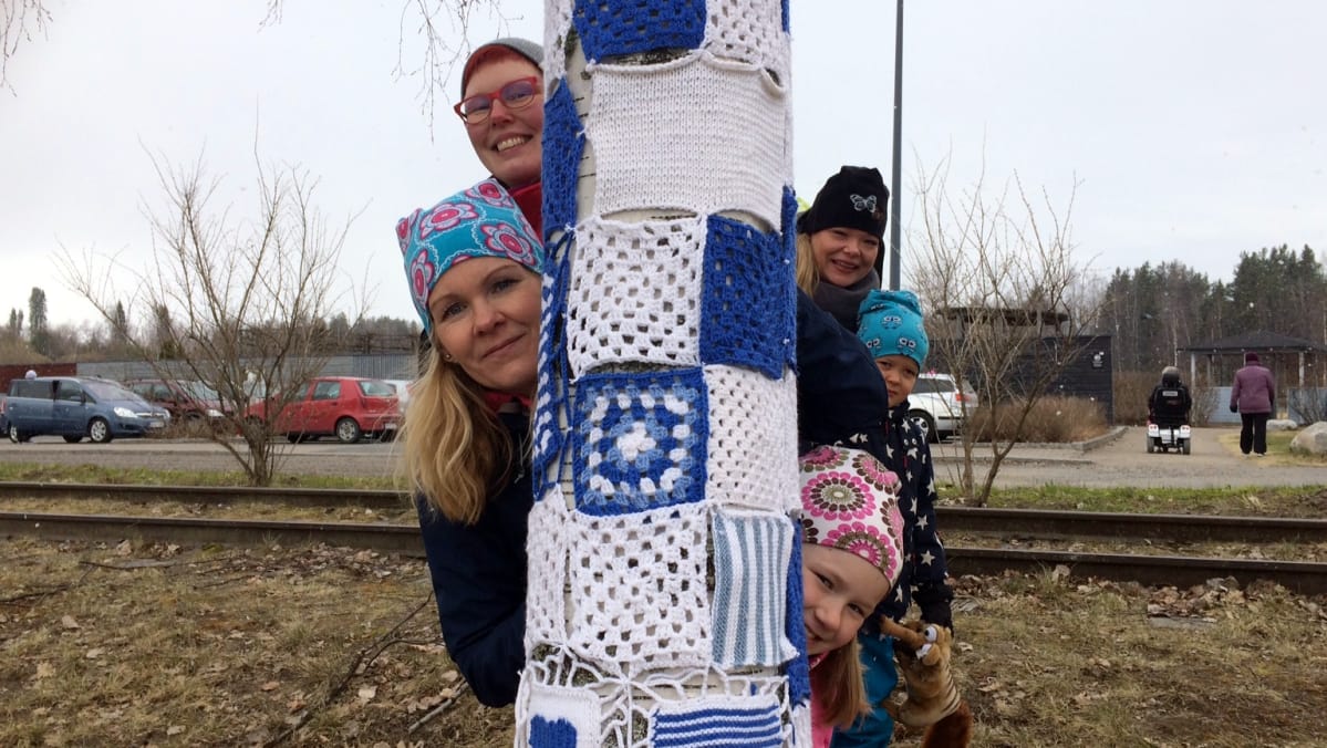 Ihmiset kurkistavat neule- ja virkkuutöin koristellun puun takaa.