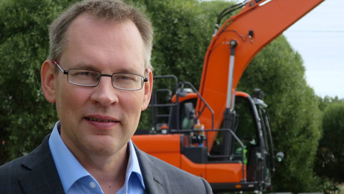 Silmälaspäinene mies seisoo kaivinkoneen edessä
