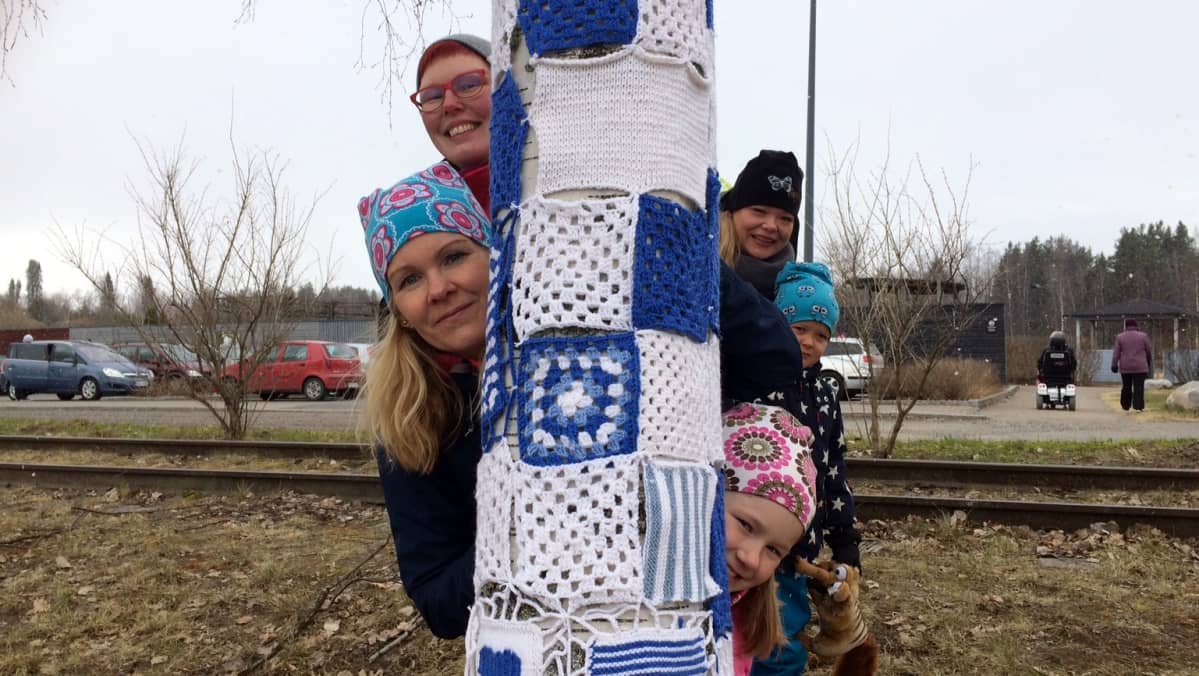 Ihmiset kurkistavat neule- ja virkkuutöin koristellun puun takaa.