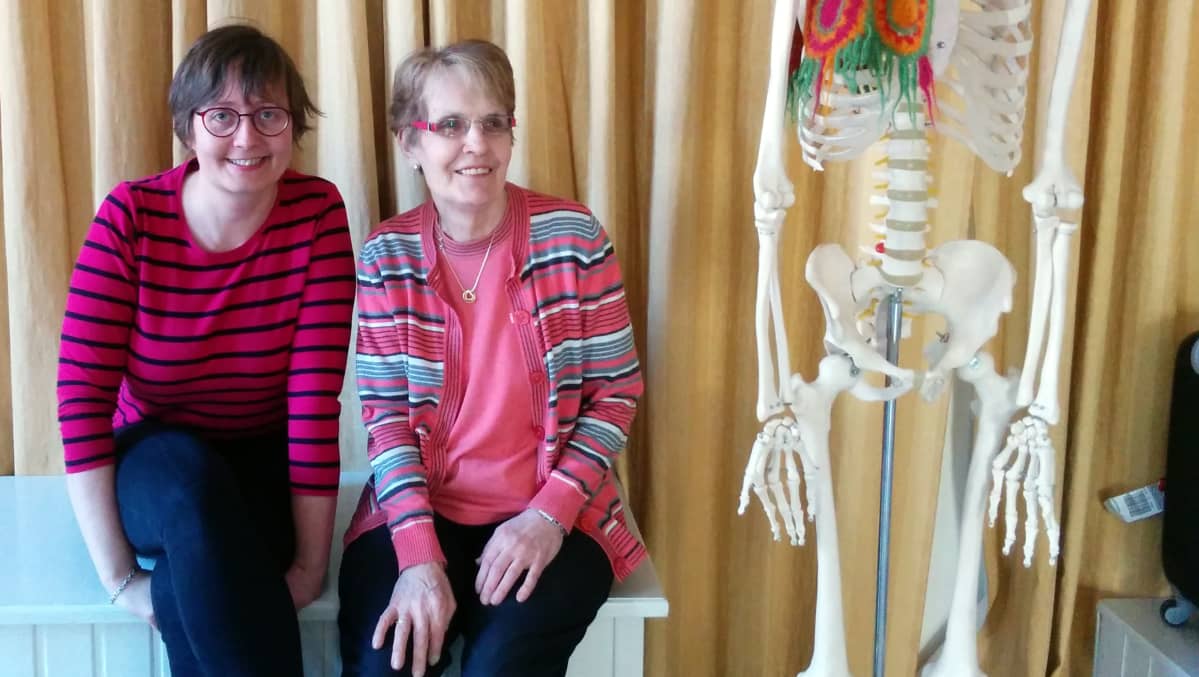 Katja Seppinen ja Pirkko Sysimetsä istuvat penkillä. Kuvan oikeassa laidassa näkyy osa muovisesta luurangosta.