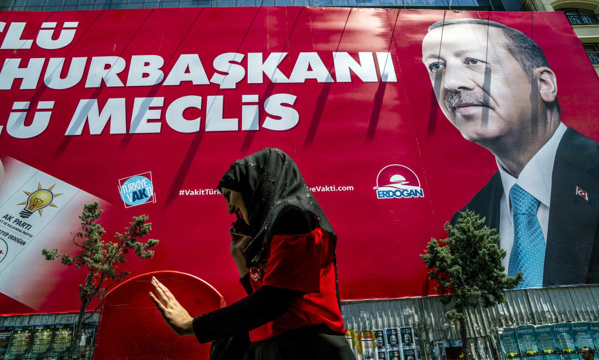 Nainen kävelee kadulla ja juttelee kännykkäänsä. Takana näkyy iso Recep Tayyip Erdoğanin punapohjainen kampanjajuliste.