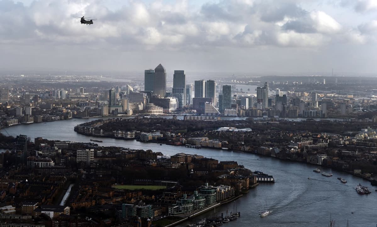Maisemakuva Lontoosta. Kuvassa keskialalla on rykelmä Itä-Lontoossa sijaitsevan Canary Wharfin finanssikeskuksen pilvenpiirtäjiä. Taivas on pilvinen. Kuvassa ylävasemmalla lentää helikopteri. Thamesjoki kiemurtelee kuvassa oikeasta alalaidasta vasemmalle, jossa se tekee mutkan ja etenee oikealle Canary Wharfin pilvenpiirtäjien editse kunnes jossain näkymättömissä taas tekee käännöksen vasempaan. Joella kulkee laivoja.