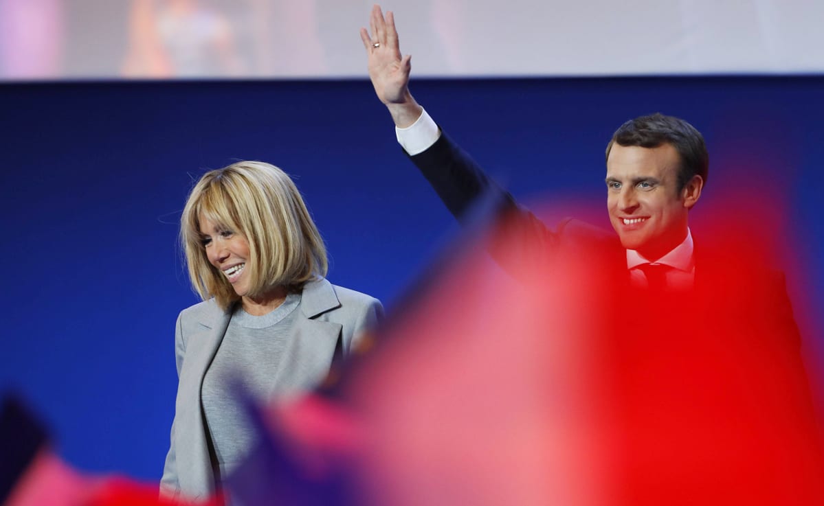 Brigitte Trogneux ja Emmanuel Macron seisovat esiintymislavalla. Macron heiluttaa kättään. Etualalla joku heiluttaa jonkinlaista punaista vaatetta, joka näkyy kuvassa punaisen värin leiskahduksena.