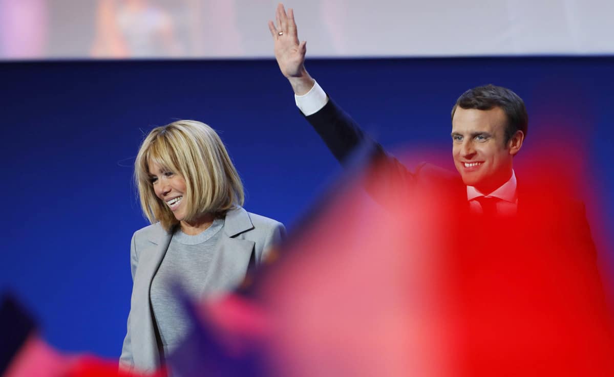 Brigitte Trogneux ja Emmanuel Macron seisovat esiintymislavalla. Macron heiluttaa kättään. Etualalla joku heiluttaa jonkinlaista punaista vaatetta, joka näkyy kuvassa punaisen värin leiskahduksena.