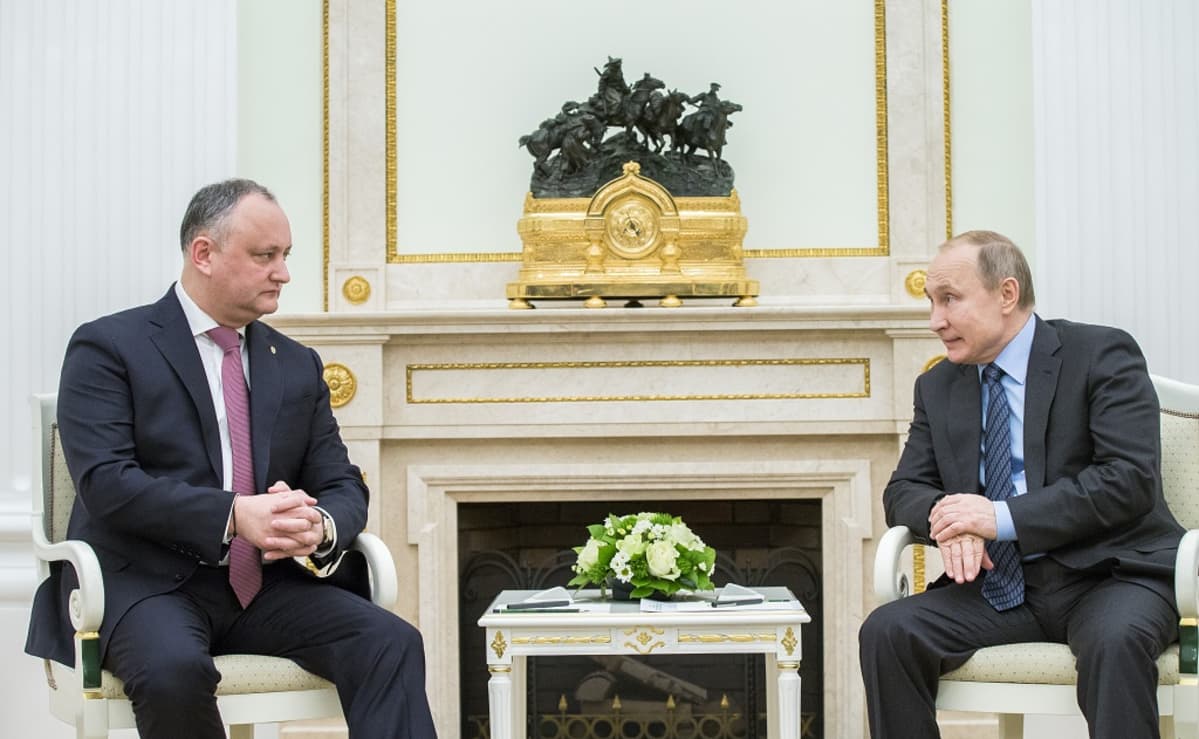 Dodon ja Putin istuvat tuoleilla takan edessä. Putin puhuu, Dodon kuuntelee. Miehillä on harmaat puvut, Putinilla sininen kravatti ja vaaleansininen kauluspaita, Dodonilla punainen kravatti. Takan reunuksella on iso kultainen kello, jonka päällä on hevoslaumaa ohjaavaa ratsumiestä esittävä patsas.