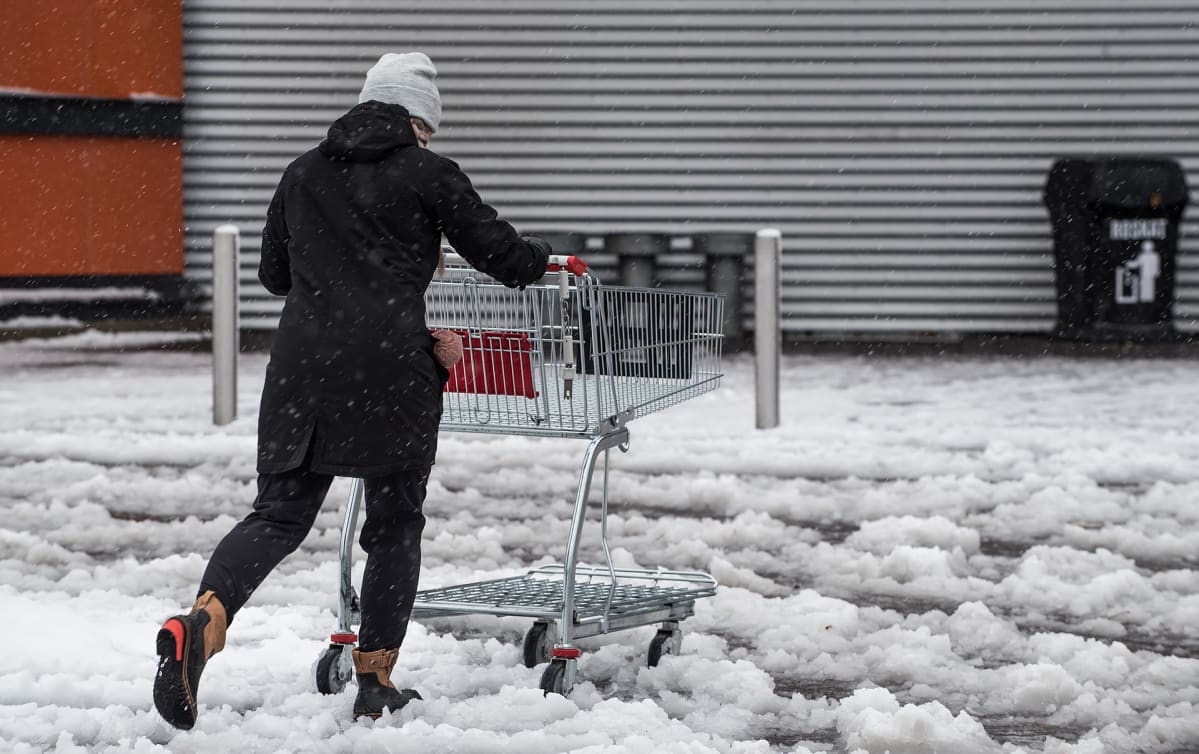 Nainen työntää ostoskärryjä lumisohjoisella parkkipaikalla.