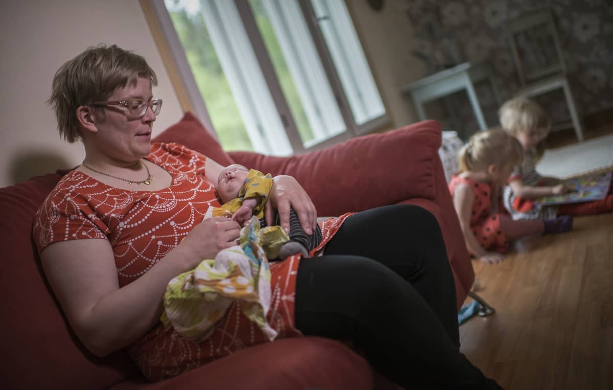 Keskosvauva Tiitus Hermanni äitinsä Suvi Toikkasen sylissä. Vanhemmat lapset lukevat satukirjaa taustalla.