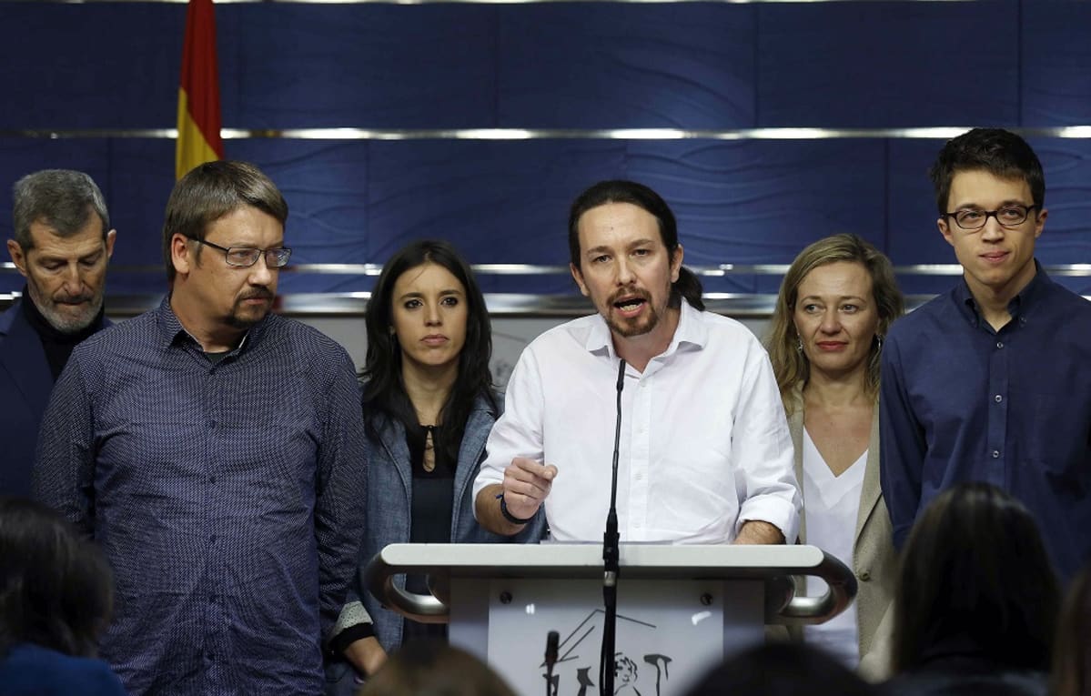 Pablo Iglesias puhuu tiedotustilaisuudessa puhujanpöntön takaa. Hänen molemmilla puolillaan seisoo joukko muita puolueen edustajia, kaksi naista ja kolme miestä. Parrakkaalla Iglesiasilla on valkoinen kauluspaita ja hänen hiuksensa on sidottu poninhännälle. Taustalla näkyy ilmeisesti Espanjan lippu.
