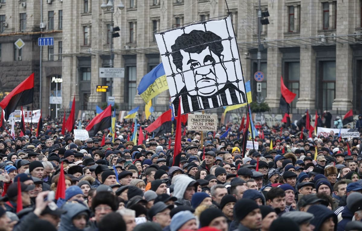 Suuri väkijoukko kulkee stalinistisen hallintorakennuksen edustalla. Kuvan keskellä näkyy suuri mustavalkoinen kyltti, johon on kuvattu karikatyyrimaisesti presidentti Petro Porošenko kaltereiden takana. Mielenosoittajat kantavat punamustia lippuja ja Ukrainan sinikeltaisia lippuja.