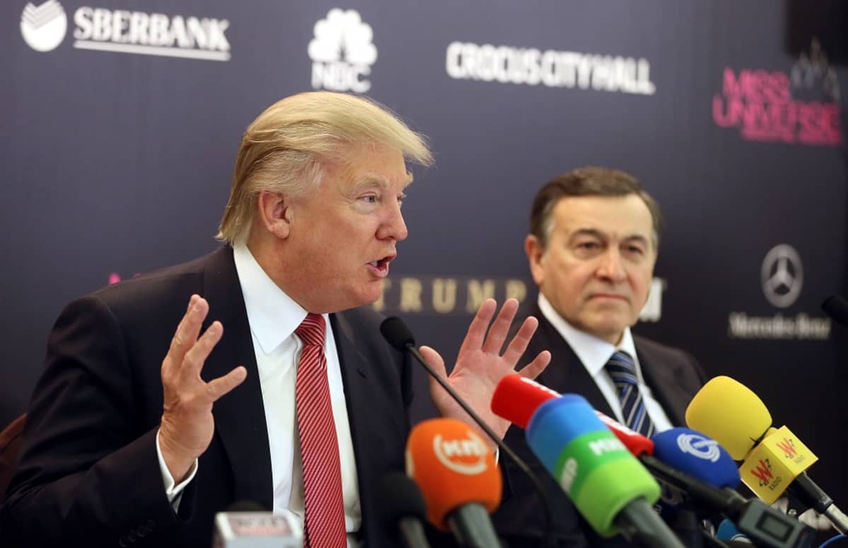 Donald Trump puhuu lehdistötilaisuudessa. Trumpin edessä on useita erivärisiä mikrofoneja. Hänen vieressään istuu Aras Agalarov. Miehet ovat tummissa puvuissa, kravatit kaulassa.