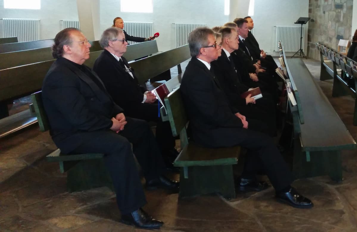 Mustiin pukeutuneet vapaaehtoiset saattavat istuvat Honkanummen Ison Kappelin pitkissä penkeissä.