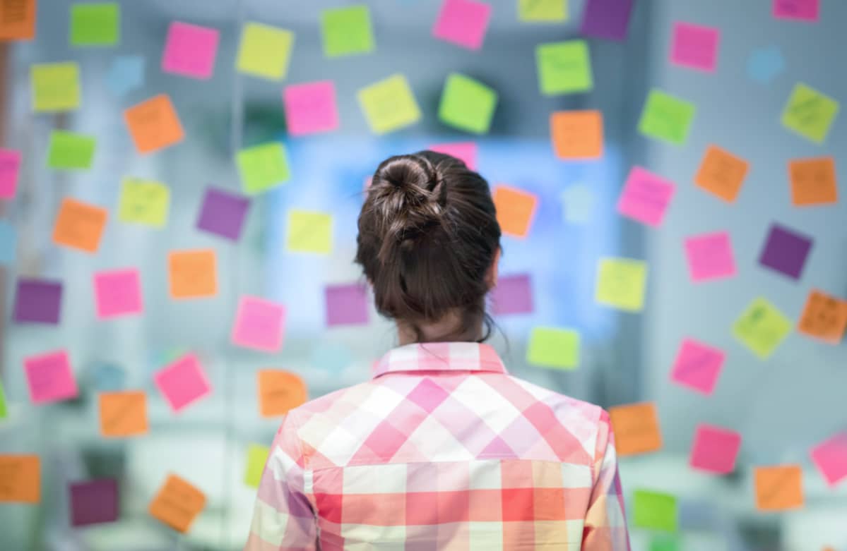 Nainen selkä kameraan päin katsoo lasiseinään kiinnitettyjä post-it-lappuja.