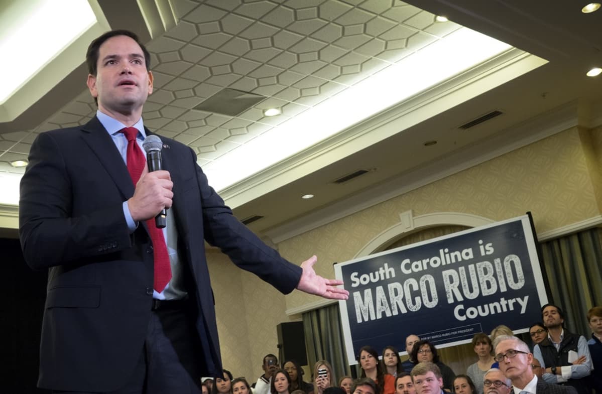 Marco Rubio puhuu hotellin salissa oikeassa kädessään olevaan mikrofoniin ja viittilöi vasemmalla kädellään takavasemmalle, jossa näkyy kampanjajuliste. Julisteessa lukee, että Etelä-Carolina on Marco Rubion maata. Taustalla istuu yleisöä. Rubiolla on tumma puku, punainen kravatti.