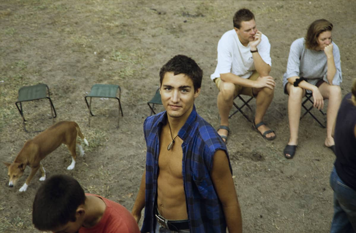 Justin Trudeau hymyilee kameralle taustallaan leirijakkaroita, joilla istuu kaksi ihmistä.