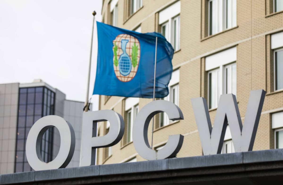 Kuvassa OPCW:n logo ja lippu, joka heiluu päämajan fasadissa.