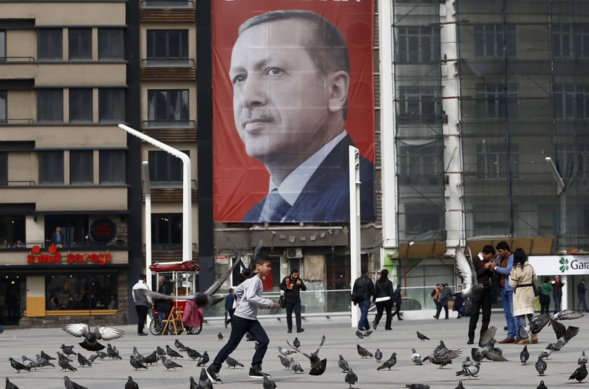 Recep Tayyip Erdoğanin jättimäinen kuva kerrostalon seinässä. Edustalla ihmisiä ja puluja.