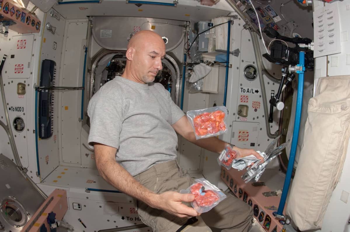 Astronautti leijailee istuma-asennossa avaruusaseman sisällä. Hänellä on käsissään useita pakkauksia pakastekuivattua ruokaa, ja kaksi pakkausta leijailee ilmassa.