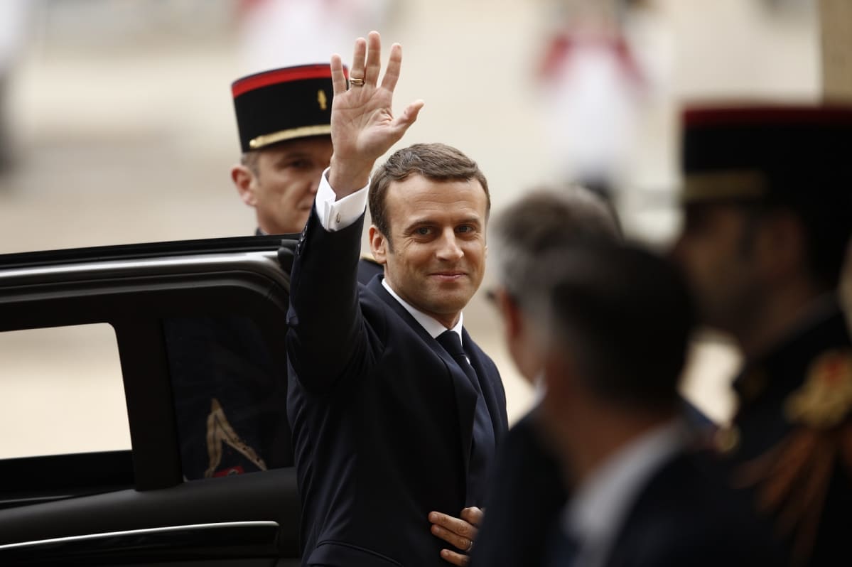 Emmanuel Macron astuu ulos autosta ja tervehtii kohti kameraa.