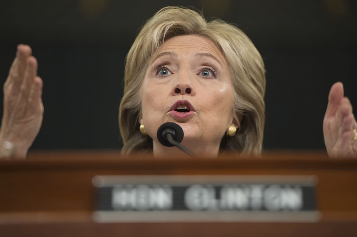 Hillary Clinton puhuu mikrofoniin ja tehostaa puhetta käsillään. Tausta on tumma. Edustalla näkyy häimeänä hänen nimikylttinsä.